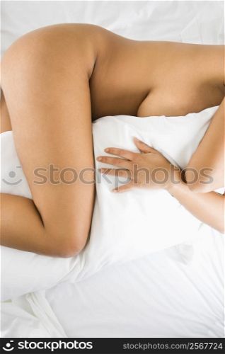 Nude woman hugging pillow lying on bed with white sheets.
