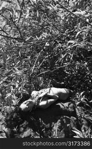 Nude young adult woman lying on back in forest with hands on chest and eyes closed.