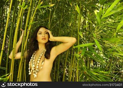 Nude young adult woman standing in bamboo with hand on head looking out.