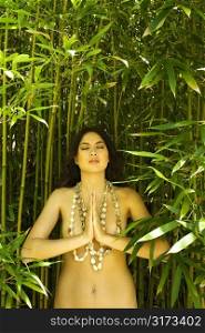 Nude young adult woman standing in bamboo with hands to chest in prayer position and eyes closed.