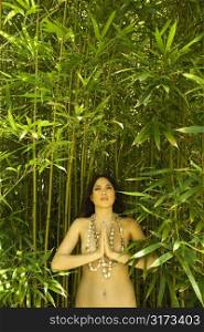 Nude young adult woman standing in bamboo with hands to chest in prayer position and looking up.