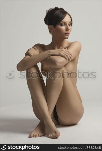 Nude, young, natural, sensual woman with brown hair and hair bun sitting on the floor holding legs near body in studio