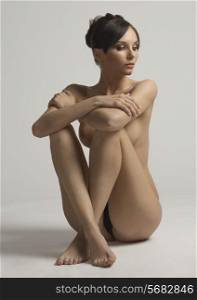 Nude, young, natural, sensual woman with brown hair and hair bun sitting on the floor holding legs near body in studio