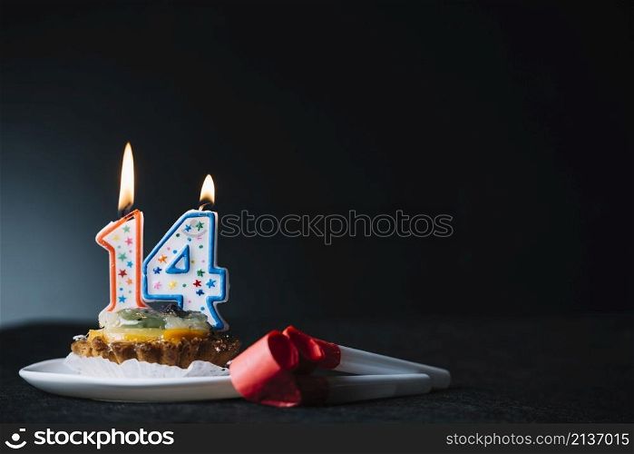 number 14 birthday lighted candle slice tart party horn blower against black backdrop