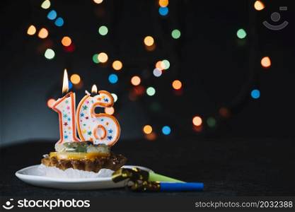 number 16 birthday lighted candle slice tart with party horn blower against illuminated bokeh backdrop