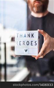 Nurse day concept. Man hands holding lightbox with Thank heroes text thanking doctors, nurses and medical staff working in hospitals during coronavirus COVID-19 pandemics. View through window glass