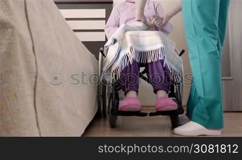 Nurse helping elder disabled woman in wheelchair to put her in bed at home or in nursing home. Slow motion