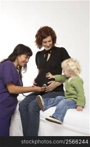 Nurse holding stethoscope on Caucasian pregnant woman&acute;s belly as daughter holds hand on belly.