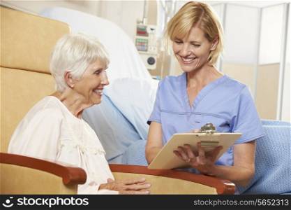Nurse Taking Notes From Senior Female Patient Seated In Chair By Hospital Bed