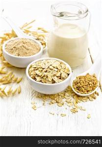Oat flakes and flour in bowls, grain in a spoon, oatmeal milk in a glass jug and oaten stalks on the background of a white wooden board