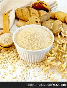Oat flour in white bowl, oat bran in spoon, oatmeal and stems, bread and biscuits on a background of wooden boards