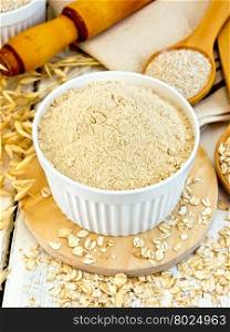 Oat flour in white bowl, oatmeal and bran in a spoon, oat stalks, a rolling pin and a napkin on a wooden boards background