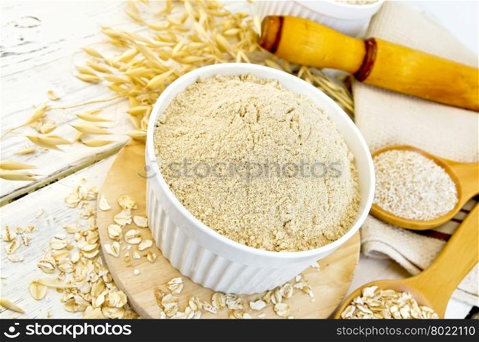 Oat flour in white bowl, oatmeal and bran in wooden spoon, stalks of oats, a rolling pin and a napkin on a wooden boards background