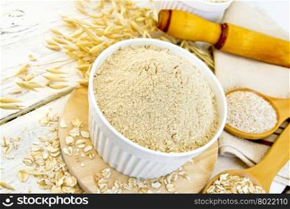 Oat flour in white bowl, oatmeal and bran in wooden spoon, stalks of oats, a rolling pin and a napkin on a wooden boards background