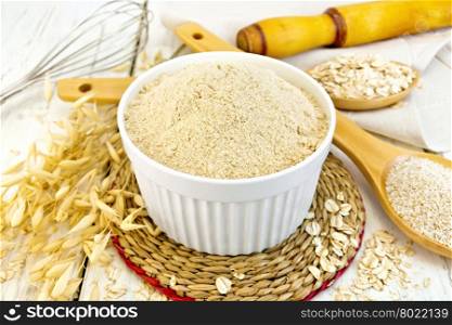 Oat flour in white bowl, oatmeal and bran in wooden spoon, stalks of oats on the background of wooden boards