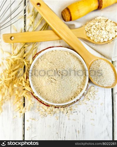 Oat flour in white bowl, oatmeal and bran in wooden spoon, stalks of oats on the background of the wooden planks on top