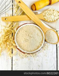 Oat flour in white bowl, oatmeal and bran in wooden spoon, stalks of oats on the background of the wooden planks on top