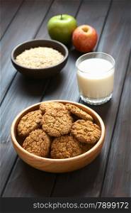 Oatmeal and apple cookies in wooden bowl with a glass of milk, apples and oatmeal in the back, photographed with natural light (Selective Focus, Focus on the front of the cookie in the middle)