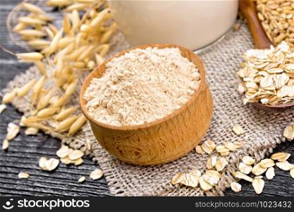 Oatmeal flour in bowl, milk in jug, oatmeal in spoon on napkin of burlap, grain in bag, oaten stalks against wooden board