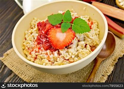 Oatmeal in a bamboo bowl with strawberry and rhubarb sauce on a napkin of burlap, spoon, milk in glass jug, rhubarb on a dark board