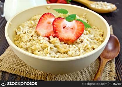 Oatmeal in a yellow cup with strawberries on a napkin of burlap, spoon, milk in a glass jar on the background of dark board
