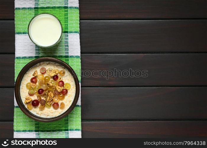 Oatmeal porridge with grapes and walnuts in rustic bowl, glass of milk above, photographed overhead on dark wood with natural light