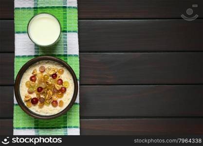 Oatmeal porridge with grapes and walnuts in rustic bowl, glass of milk above, photographed overhead on dark wood with natural light