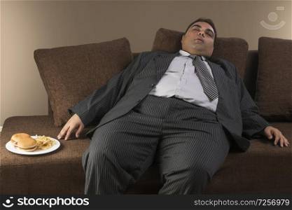 Obese businessman in formal clothes sleeping on sofa with plate of burgers and french fries