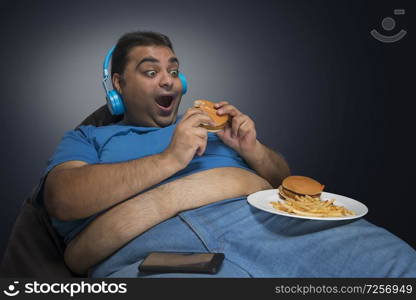 Obese man sitting on bean bag enjoying burgers and french fries listening to music on headphones