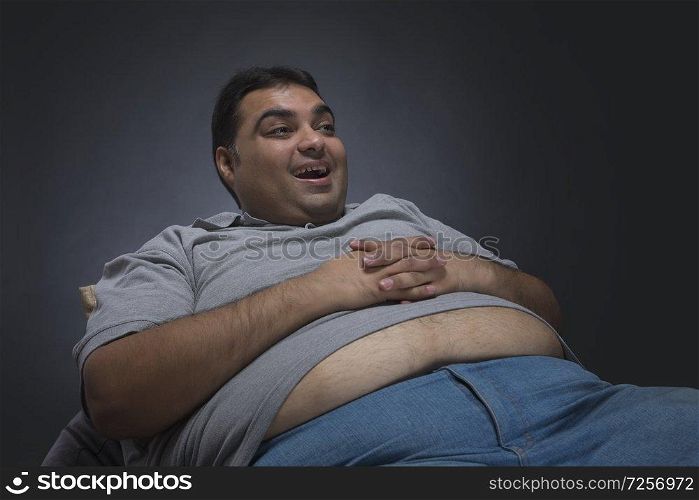 Obese man with hands on his belly visible out of his shirt sitting on a chair looking away and smiling