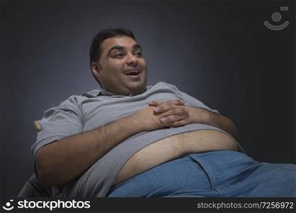 Obese man with hands on his belly visible out of his shirt sitting on a chair looking away and smiling
