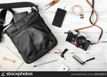 Objects on wooden background: leather bag, camera, smartphone, keys, knife. Outfit of young man.