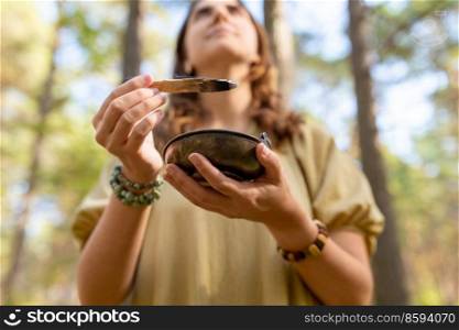 occult science and supernatural concept - close up of woman or witch with smoking palo santo stick and bowl performing magic ritual in forest. woman with palo santo performing magic ritual
