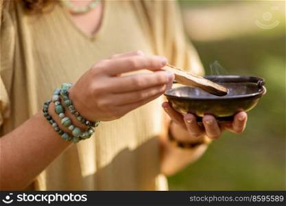 occult science and supernatural concept - close up of woman or witch with smoking palo santo stick and bowl performing magic ritual in forest. woman with palo santo performing magic ritual