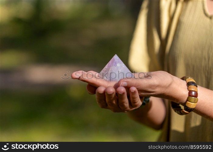 occult science and supernatural concept - close up of woman or witch with semiprecious crystal or gemstone pyramid performing magic ritual in forest. close up of hands holding crystal pyramid