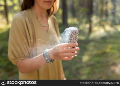 occult science and supernatural concept - close up of woman or witch with smoking white sage performing magic ritual in forest. woman with sage performing magic ritual in forest