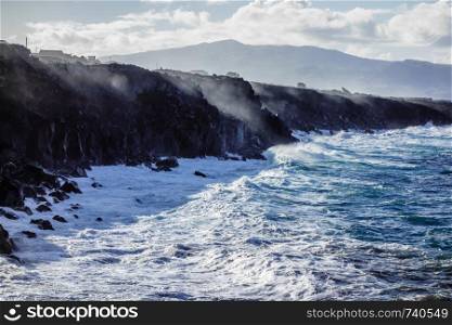 Ocean waves splashing on jagged cliffs with spray reflecting sunlight, in Azores, Portugal.
