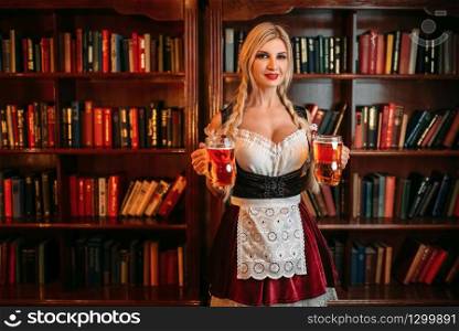 Octoberfest waitress with two mugs of fresh beer standing against shelf with books in vintage pub. Sexy barmaid with attractive shapes in traditional style dress holds glass of foamy beverage