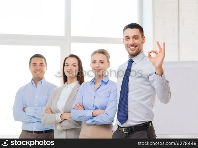 office, business, and teamwork concept - friendly young smiling businessman with team on back showing ok-sign