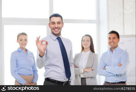 office, business, and teamwork concept - friendly young smiling businessman with team on back showing ok-sign