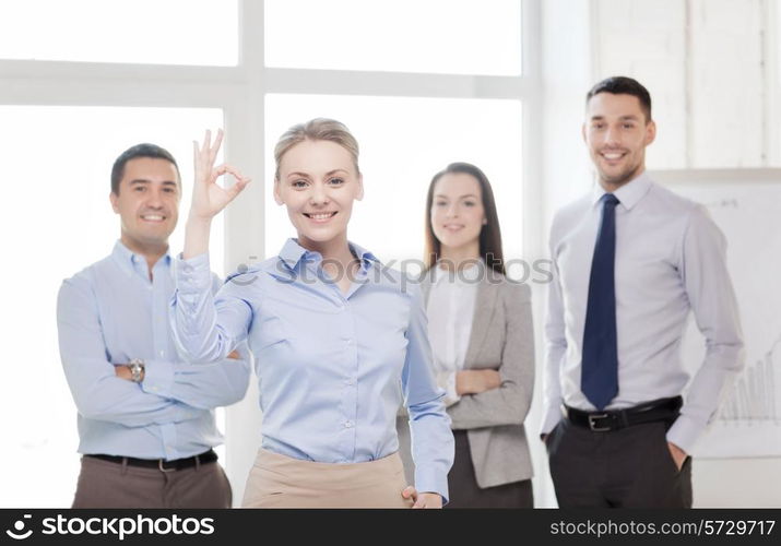 office, business and teamwork concept - friendly young smiling businesswoman with team on back showing ok-sign