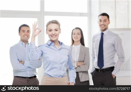 office, business and teamwork concept - friendly young smiling businesswoman with team on back showing ok-sign
