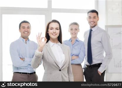 office, business and teamwork concept - friendly young smiling businesswoman with team on back showing ok-sign