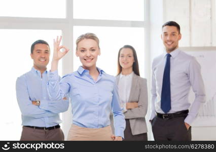 office, business and teamwork concept - friendly young smiling businesswoman with team on back showing ok-sign