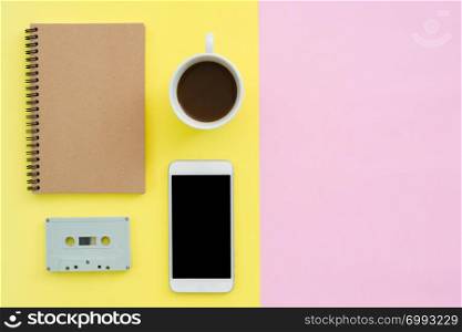 Office desk working space - Flat lay top view of working space with white blank notebook, cassette, coffee cup and mock up phone on pastel background. Pastel pink yellow color background space concept