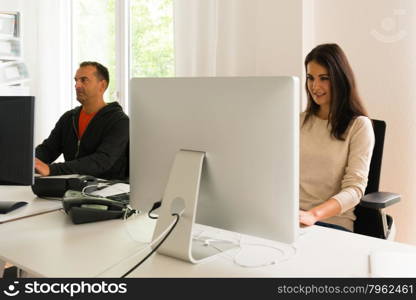 Office people being absorbed in their work. Office people being absorbed in their work: Attractive young woman working in front of her modern bright computer screen with headphones on her table and older guy sitting in front of his screen and telephone next to her.