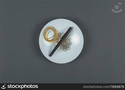 Office stationery supplies including a pen, rubber bands and paper clips on a white ceramic plate in a conceptual image, viewed from above centered on a blue background with copyspace