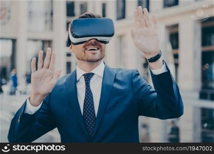 Office worker in suit on his break exploring virtual reality with VR glasses, sitting outside with his hands in air, people and buildings in blurred background, businessman enjoying gaming experience. Office worker in formal suit exploring virtual world with VR glasses while relaxing outdoors