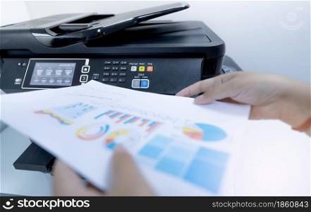 Office worker prints paper on multifunction laser printer. Copy, print, scan, and fax machine in office. Document and paper work. Print technology. Blurred hand hold printed paper. Scanner equipment.