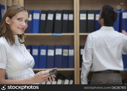 Office worker using palm top standing in file storage room, colleague standing in background
